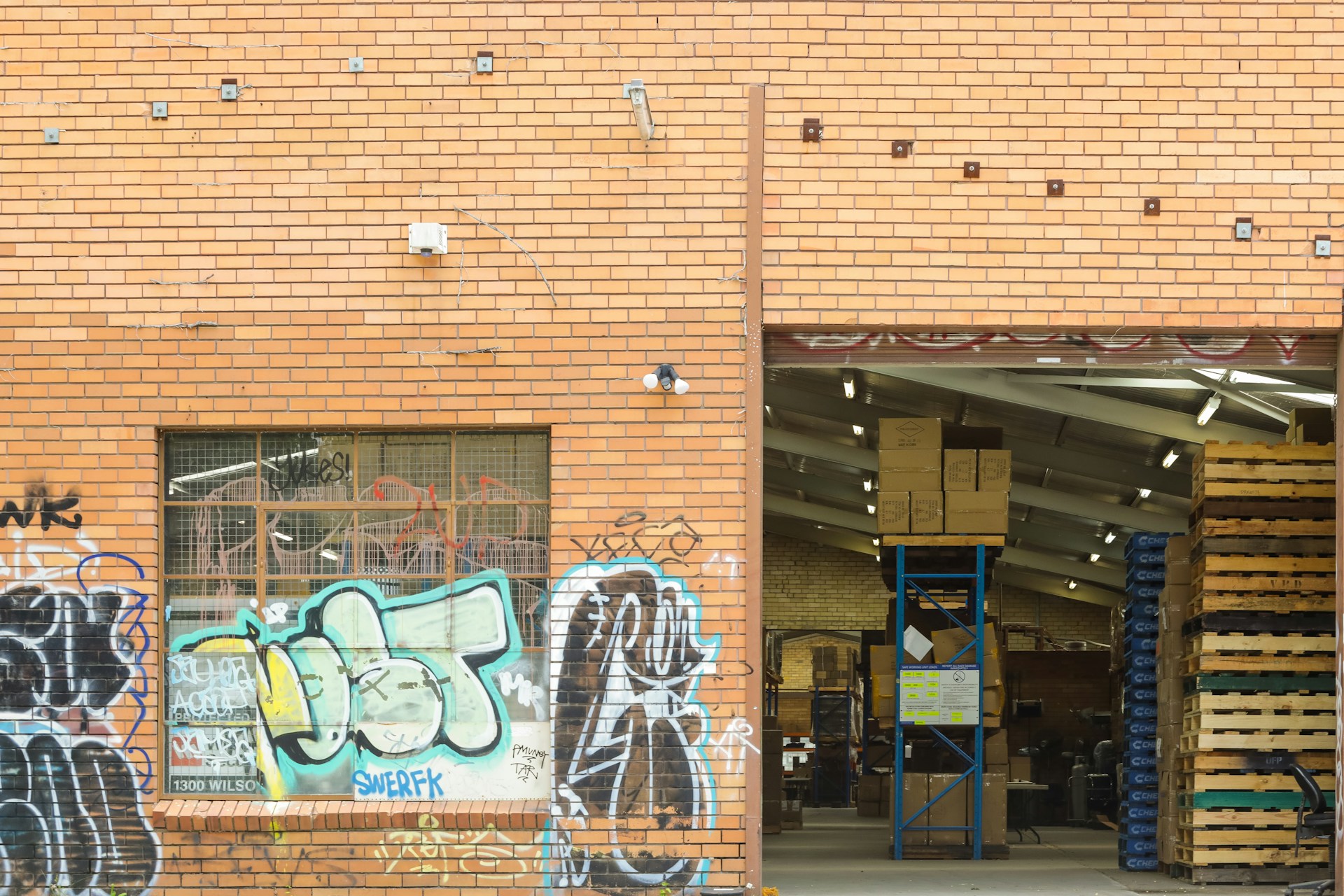 A brick warehouse exterior featuring vibrant graffiti on the walls. On the left, large graffiti designs in various colors including black, blue, and green cover the wall around a barred window. The open entrance on the right reveals an interior with stacked cardboard boxes, wooden pallets, and industrial shelving under fluorescent lights.