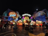 A vibrant amusement park scene featuring the entrance to a ride themed around 'The Simpsons' television show. The main feature is a large animated character head, illuminated by multicolored lights against the night sky. The area is bustling with visitors walking around, and the colorful facades of the attractions add to the lively atmosphere.