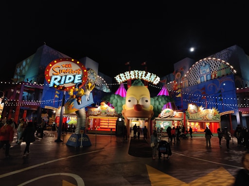 A vibrant amusement park scene featuring the entrance to a ride themed around 'The Simpsons' television show. The main feature is a large animated character head, illuminated by multicolored lights against the night sky. The area is bustling with visitors walking around, and the colorful facades of the attractions add to the lively atmosphere.