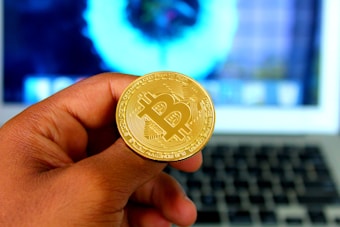 A hand holding a golden coin with the Bitcoin symbol in front of a blurred laptop screen. The focus is on the coin, emphasizing the cryptocurrency theme.