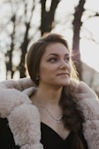 A woman with long hair is wearing a luxurious fur-lined coat and a delicate necklace. She is outdoors, with sunlight softly illuminating her face. The background consists of tall trees with blurred branches, creating a serene, natural setting.