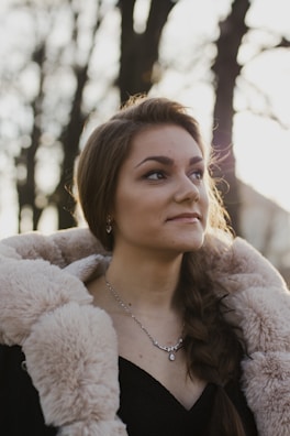 A woman with long hair is wearing a luxurious fur-lined coat and a delicate necklace. She is outdoors, with sunlight softly illuminating her face. The background consists of tall trees with blurred branches, creating a serene, natural setting.