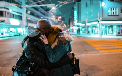 A couple exchanging warm embraces in a city street lit by twinkling evening lights.
