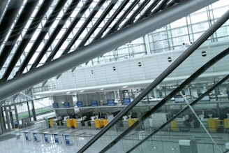 An indoor view of a modern airport terminal featuring empty check-in counters. The architecture includes sleek, diagonal metal beams and large windows allowing natural light to flood the area. Digital screens are visible above the counters, which are partitioned by metal barriers. The overall appearance is clean and minimalistic.