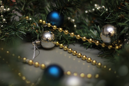 A close-up of Christmas decorations featuring green artificial pine branches, golden bead garlands, and shiny baubles in blue and silver. The arrangement is festive and appears to be part of a holiday setup.