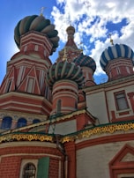 Colorful onion-shaped domes with intricate patterns and crosses on top. The building exhibits classic Russian architectural style with detailed brickwork and decorative trim. A bright blue sky with scattered clouds provides a vivid backdrop.
