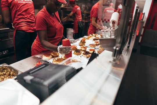 Employees in a fast food restaurant are preparing burgers at a stainless steel counter. The workers are wearing red uniforms and hats, and one of them is applying condiments to the burgers using a squeeze bottle.