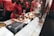 Employees in a fast food restaurant are preparing burgers at a stainless steel counter. The workers are wearing red uniforms and hats, and one of them is applying condiments to the burgers using a squeeze bottle.