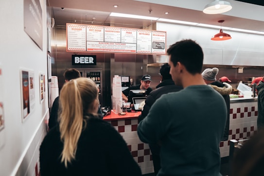 A group of people are standing in line at a counter inside a fast food restaurant. The menu board above displays various items such as burgers and hot dogs, and a neon sign labeled 'BEER' is visible. The interior is decorated with white and red tiles, and the staff member behind the counter is taking orders.