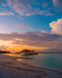 Overwater bungalows are situated on a serene beach during sunset. The sky displays a gradient of warm orange and pink hues blending into cool blues as clouds scatter across it. The calm turquoise water reflects the setting sun, creating a peaceful ambiance.