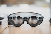 A pair of sleek, modern augmented reality glasses with large round lenses and multiple small cameras on the frame, placed on a wooden table. The design is minimalistic and high-tech, with a focus on functionality and innovation.