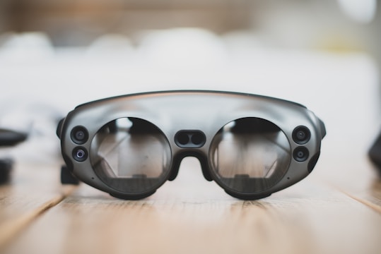 A pair of sleek, modern augmented reality glasses with large round lenses and multiple small cameras on the frame, placed on a wooden table. The design is minimalistic and high-tech, with a focus on functionality and innovation.