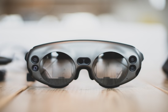 A pair of sleek, modern augmented reality glasses with large round lenses and multiple small cameras on the frame, placed on a wooden table. The design is minimalistic and high-tech, with a focus on functionality and innovation.