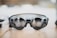 A pair of sleek, modern augmented reality glasses with large round lenses and multiple small cameras on the frame, placed on a wooden table. The design is minimalistic and high-tech, with a focus on functionality and innovation.