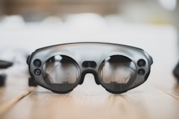 A pair of sleek, modern augmented reality glasses with large round lenses and multiple small cameras on the frame, placed on a wooden table. The design is minimalistic and high-tech, with a focus on functionality and innovation.