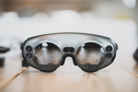 A pair of sleek, modern augmented reality glasses with large round lenses and multiple small cameras on the frame, placed on a wooden table. The design is minimalistic and high-tech, with a focus on functionality and innovation.