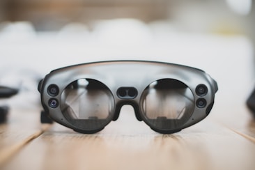 A pair of sleek, modern augmented reality glasses with large round lenses and multiple small cameras on the frame, placed on a wooden table. The design is minimalistic and high-tech, with a focus on functionality and innovation.
