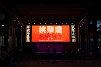 A dimly lit stage is set up with a large digital screen displaying text and some figures standing in a line. The screen is predominantly red, surrounded by illuminated panels with additional text. A group of people stand in front of the stage, possibly spectators.