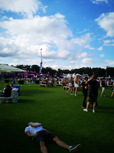 Groups of friends playing lawn games on the grass, with festival tents and colorful flags fluttering above.