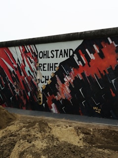 A mural on a wall features bold, abstract patterns in red, black, and white with a chain painted across it. Partial words, 'WOHLSTAND FREIHEIT' are visible amid the artwork. The ground in front appears to be disturbed soil.