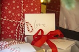 A close-up view of holiday-themed presents, featuring red wrapping paper with white snowflake patterns and white twine. An envelope with the word 'love' written in glittery letters is placed on top. A red ribbon is tied around another present, and additional cards are visible in the background.