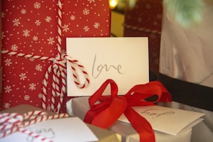 A close-up view of holiday-themed presents, featuring red wrapping paper with white snowflake patterns and white twine. An envelope with the word 'love' written in glittery letters is placed on top. A red ribbon is tied around another present, and additional cards are visible in the background.