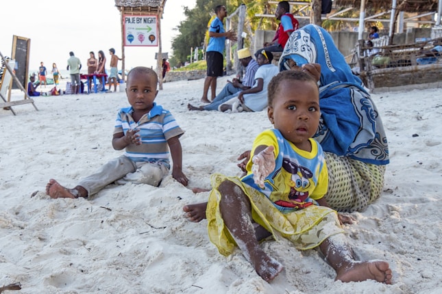 Young children are playing on a sandy beach, with one child closer to the camera reaching out. A woman in a blue headscarf and patterned clothing is seated next to them. In the background, several other people are visible, some standing and others sitting. There is a signboard that reads 'PAJE BY NIGHT' and a few shaded structures with wooden frameworks.
