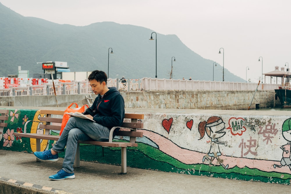 man reading book while sitting on bench beside wall with mural during day