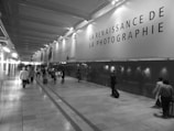 A hallway with a high ceiling is lined by a large advertising panel displaying a message about the rebirth of photography. There are several people walking, some with luggage, suggesting an airport or train station setting. The image is in black and white, giving it a classic or timeless feel.