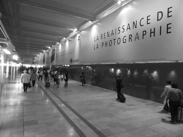 A hallway with a high ceiling is lined by a large advertising panel displaying a message about the rebirth of photography. There are several people walking, some with luggage, suggesting an airport or train station setting. The image is in black and white, giving it a classic or timeless feel.