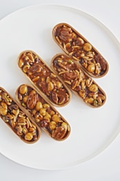 Five elongated pastries filled with a variety of nuts and seeds, drenched in a glossy caramelized coating, are arranged on a clean, white plate. The nuts and seeds include almonds, hazelnuts, and pistachios, contributing to a rich texture and appearance.