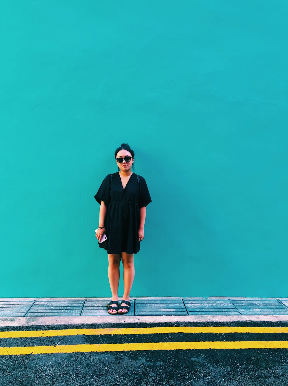 Mujer con vestido negro mientras sostiene un teléfono inteligente y está de pie junto a la pared verde azulado