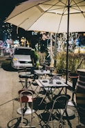 Tables and chairs are set up outdoors on a city sidewalk under a large white patio umbrella. Several chairs are arranged around foldable tables, some with plates and small candle holders. A car is parked in the background, and the scene is illuminated by streetlights and nearby building lights, creating a cozy evening atmosphere.
