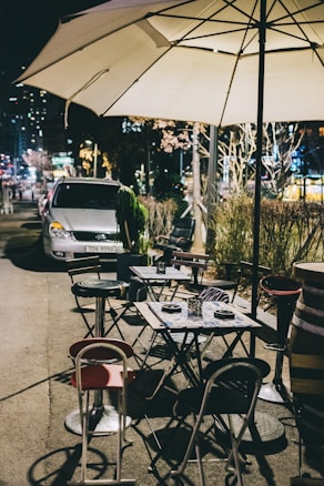 Tables and chairs are set up outdoors on a city sidewalk under a large white patio umbrella. Several chairs are arranged around foldable tables, some with plates and small candle holders. A car is parked in the background, and the scene is illuminated by streetlights and nearby building lights, creating a cozy evening atmosphere.