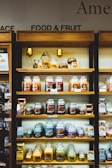 Shelves filled with a variety of scented candles in glass jars, each featuring different labels and colors. The display is neatly organized on wooden shelves and includes additional small decorative items like miniature pumpkins. Each shelf showcases different candle collections, arranged side by side.