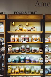 Shelves filled with a variety of scented candles in glass jars, each featuring different labels and colors. The display is neatly organized on wooden shelves and includes additional small decorative items like miniature pumpkins. Each shelf showcases different candle collections, arranged side by side.