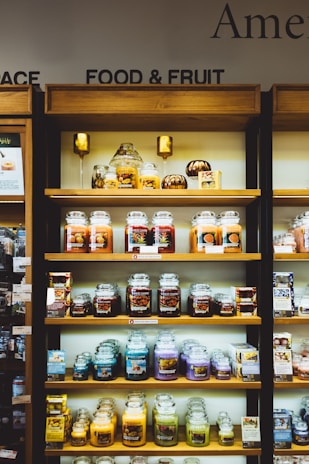 Shelves filled with a variety of scented candles in glass jars, each featuring different labels and colors. The display is neatly organized on wooden shelves and includes additional small decorative items like miniature pumpkins. Each shelf showcases different candle collections, arranged side by side.