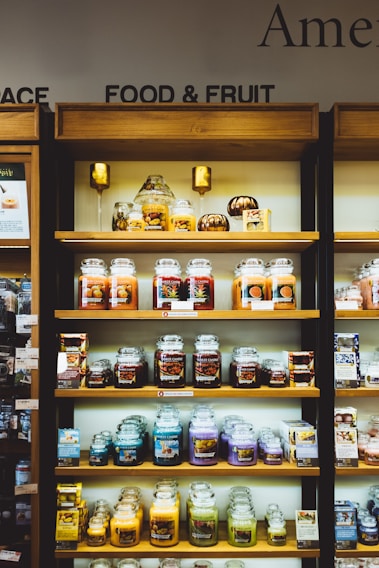 Shelves filled with a variety of scented candles in glass jars, each featuring different labels and colors. The display is neatly organized on wooden shelves and includes additional small decorative items like miniature pumpkins. Each shelf showcases different candle collections, arranged side by side.