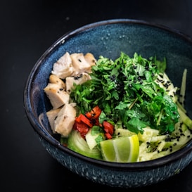 A vibrant and fresh bowl of food with cooked chicken pieces, a medley of greens, red bell peppers, lime wedges, cucumbers, and garnished with black sesame seeds, all arranged in a dark blue bowl on a black surface.