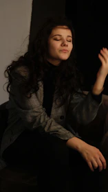 A person with long curly hair is sitting with eyes closed, appearing relaxed. They are wearing a patterned blazer over a dark top. The background is mostly dark, adding to the intimate atmosphere.