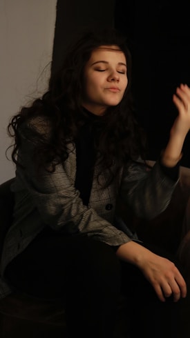A person with long curly hair is sitting with eyes closed, appearing relaxed. They are wearing a patterned blazer over a dark top. The background is mostly dark, adding to the intimate atmosphere.