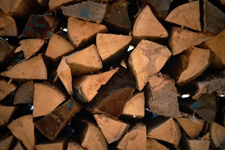 A close-up view of a neatly stacked pile of chopped firewood. The logs are cut into triangular and polygonal shapes, showing various textures and grain patterns. Some have darkened edges or markings, indicating aging or exposure to elements.