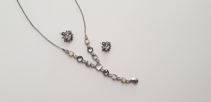 A delicate and elegant jewelry set consisting of a necklace and a pair of matching earrings is displayed against a plain light background. The necklace features a series of round and square gemstones in shades of silver and light yellow, arranged along a thin chain. The earrings are intricate clusters of similarly colored stones.