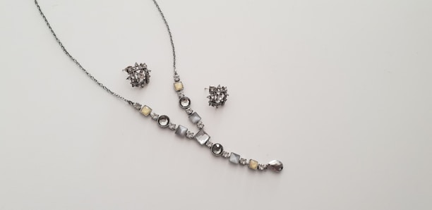 A delicate and elegant jewelry set consisting of a necklace and a pair of matching earrings is displayed against a plain light background. The necklace features a series of round and square gemstones in shades of silver and light yellow, arranged along a thin chain. The earrings are intricate clusters of similarly colored stones.