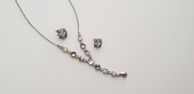 A delicate and elegant jewelry set consisting of a necklace and a pair of matching earrings is displayed against a plain light background. The necklace features a series of round and square gemstones in shades of silver and light yellow, arranged along a thin chain. The earrings are intricate clusters of similarly colored stones.