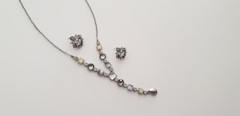 A delicate and elegant jewelry set consisting of a necklace and a pair of matching earrings is displayed against a plain light background. The necklace features a series of round and square gemstones in shades of silver and light yellow, arranged along a thin chain. The earrings are intricate clusters of similarly colored stones.