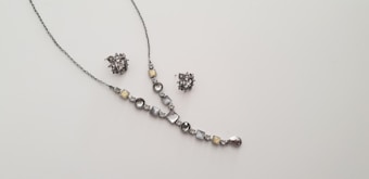 A delicate and elegant jewelry set consisting of a necklace and a pair of matching earrings is displayed against a plain light background. The necklace features a series of round and square gemstones in shades of silver and light yellow, arranged along a thin chain. The earrings are intricate clusters of similarly colored stones.