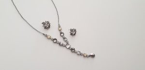 A delicate and elegant jewelry set consisting of a necklace and a pair of matching earrings is displayed against a plain light background. The necklace features a series of round and square gemstones in shades of silver and light yellow, arranged along a thin chain. The earrings are intricate clusters of similarly colored stones.