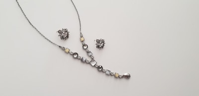 A delicate and elegant jewelry set consisting of a necklace and a pair of matching earrings is displayed against a plain light background. The necklace features a series of round and square gemstones in shades of silver and light yellow, arranged along a thin chain. The earrings are intricate clusters of similarly colored stones.