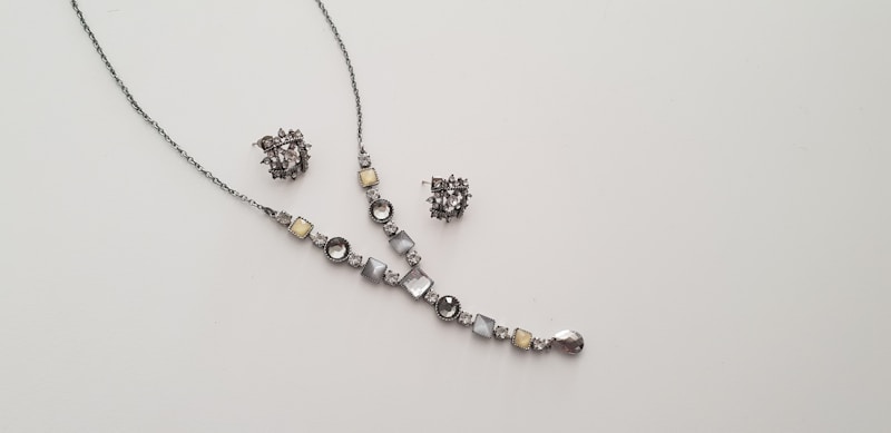 A delicate and elegant jewelry set consisting of a necklace and a pair of matching earrings is displayed against a plain light background. The necklace features a series of round and square gemstones in shades of silver and light yellow, arranged along a thin chain. The earrings are intricate clusters of similarly colored stones.